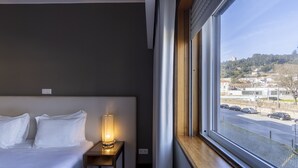 A hotel room with a bed, a bedside table with a lamp, and a window with a view of a street and buildings.