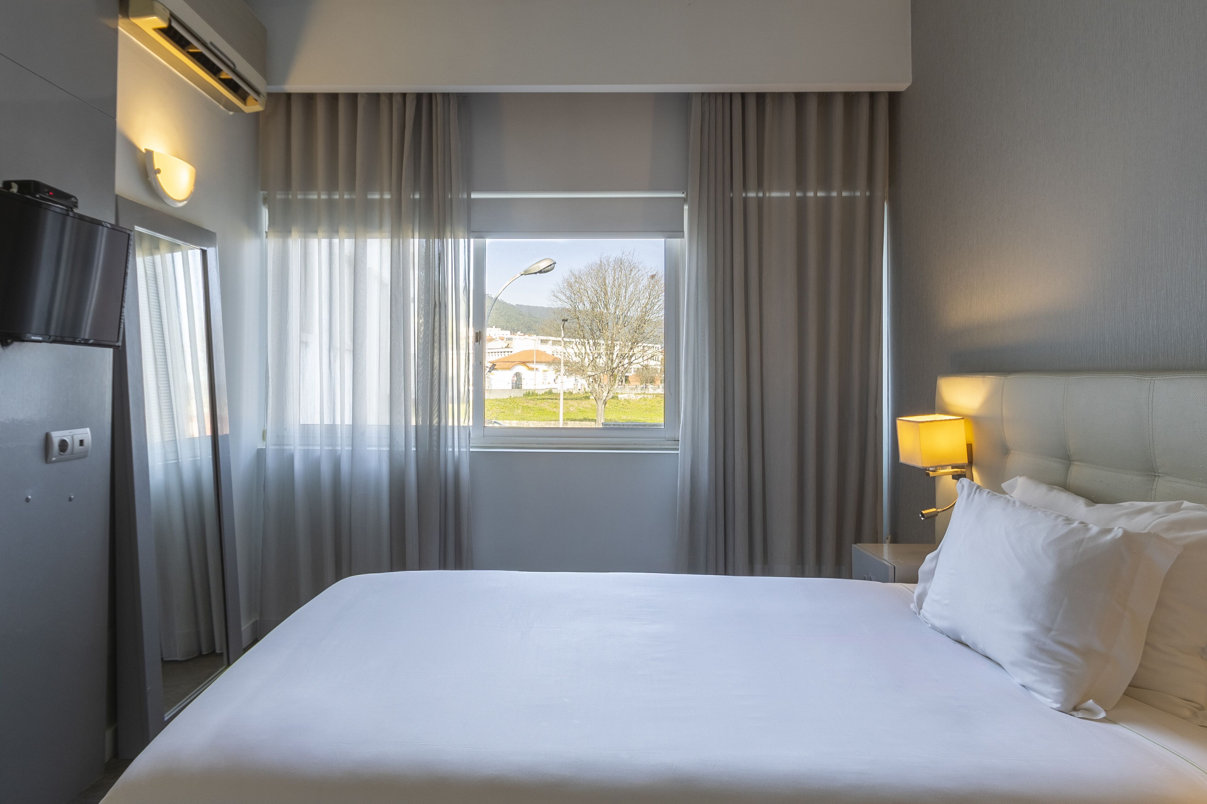 A hotel room with a bed, curtains, a window with a view, a wall-mounted TV, and a bedside lamp.