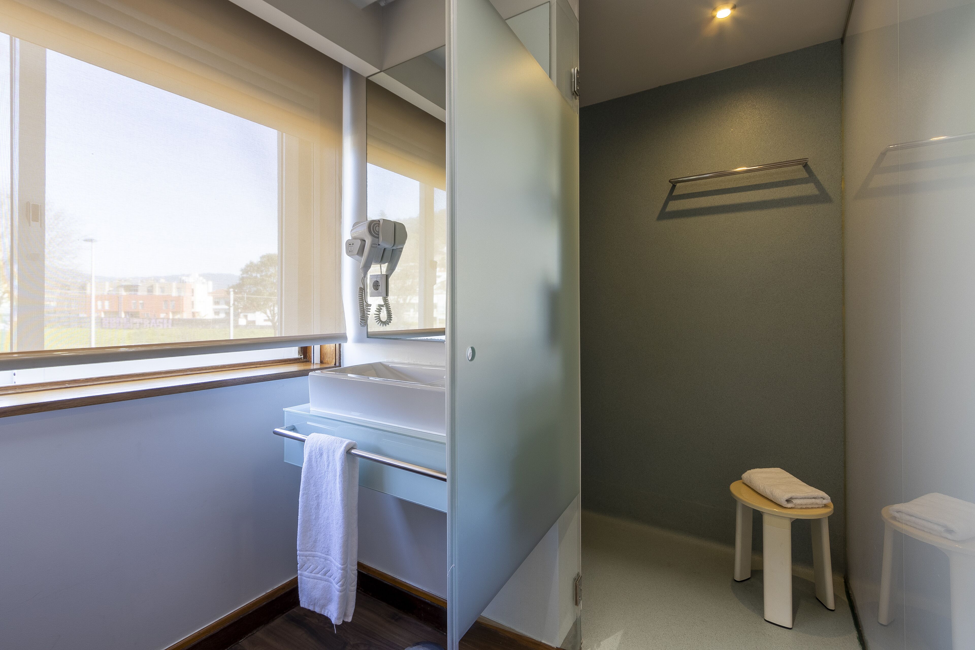A modern bathroom with a window, a hairdryer, a towel rack, and a small stool.