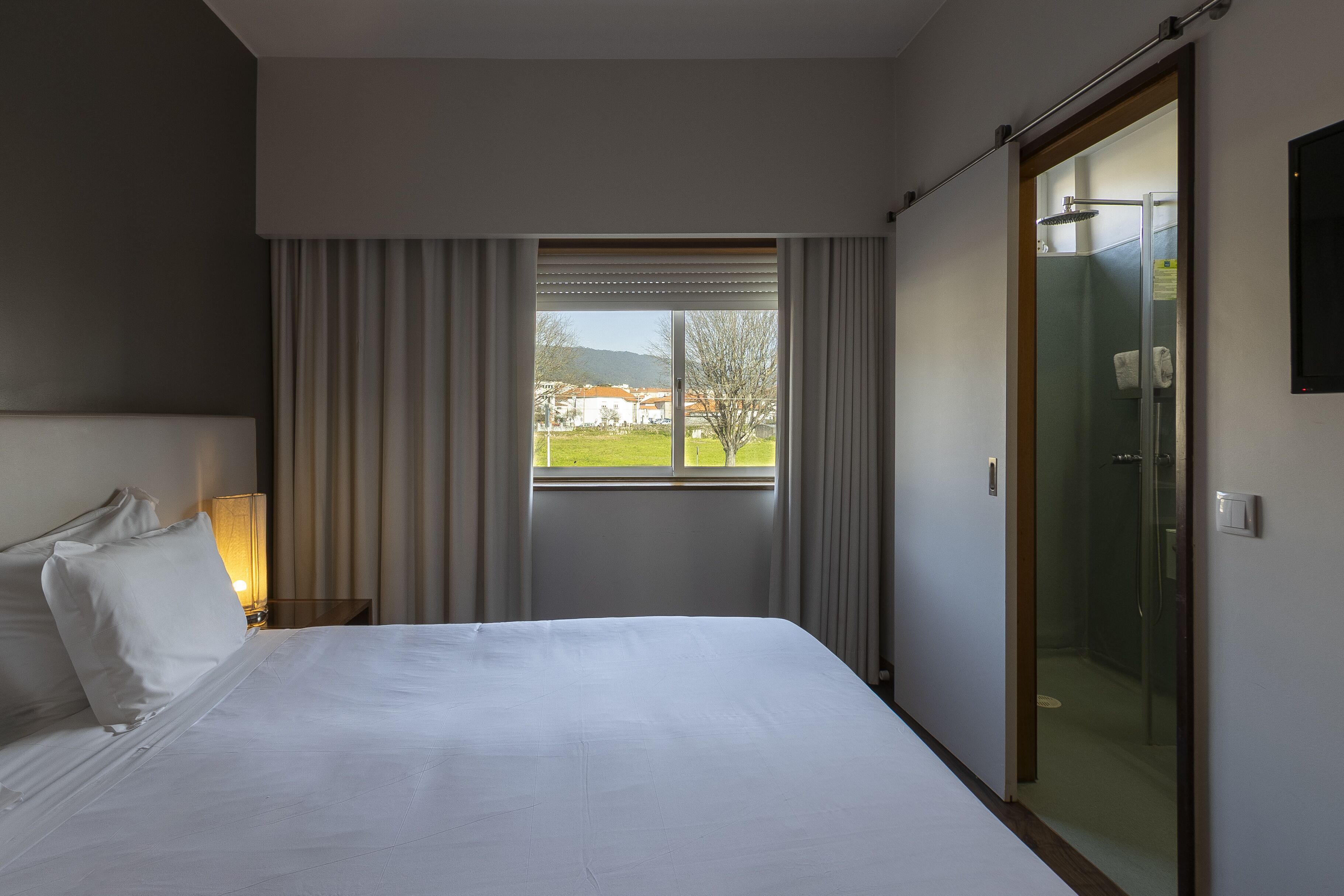 A hotel room with a bed, a window with curtains, a nightstand with a lamp, and a glass shower door.