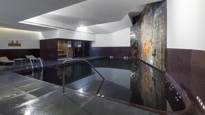 A modern indoor swimming pool with a glass wall, a seating area, and a mural on the wall.