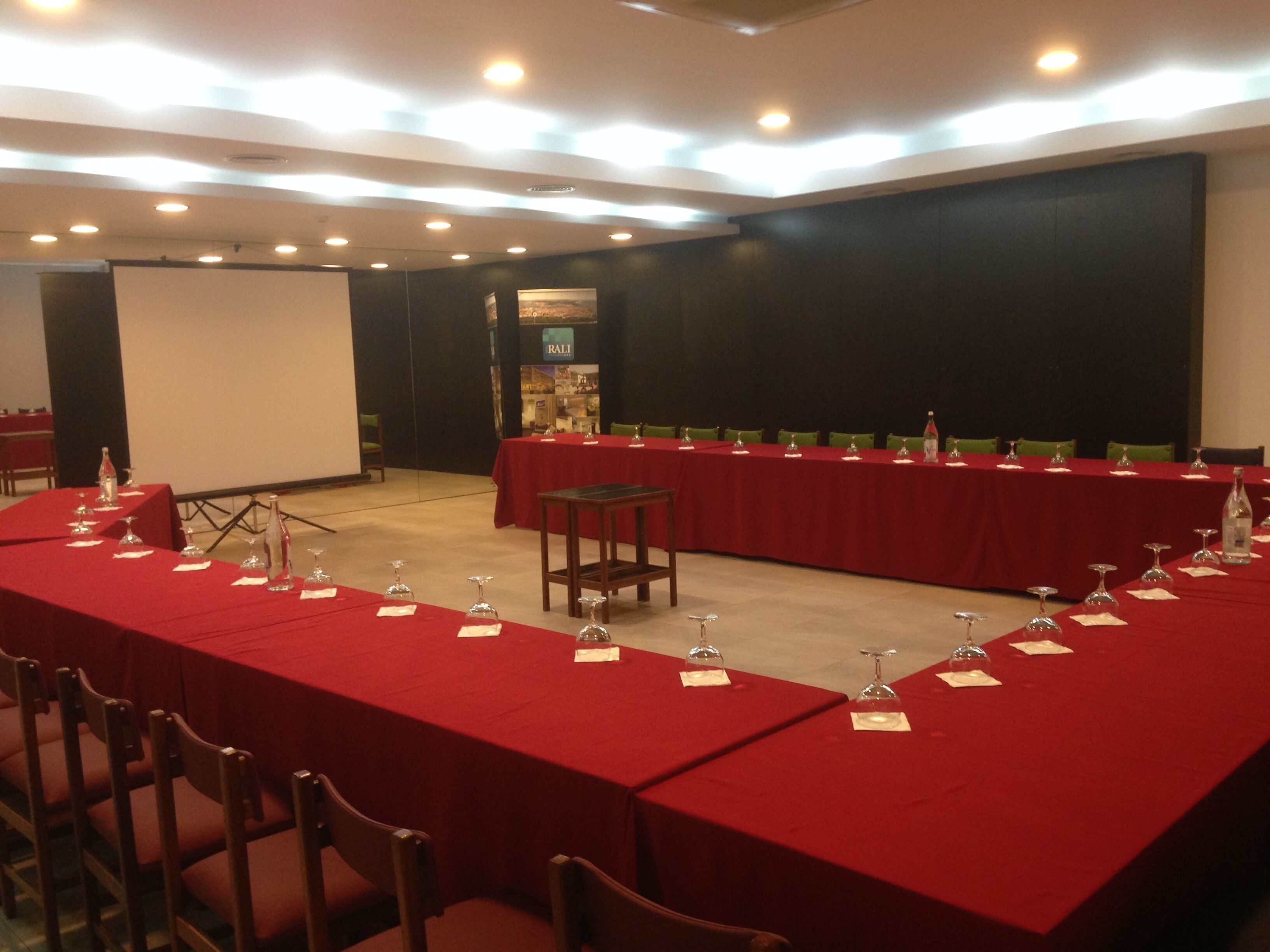 Un salón de conferencias con mesas largas cubiertas de manteles rojos, equipado con copas de vino y botellas de agua.