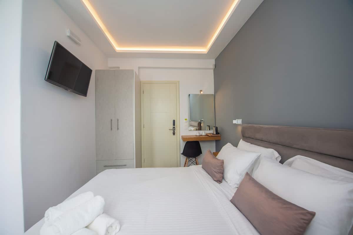 A modern hotel room with a bed, a flat-screen TV, a wardrobe, a desk with a mirror, and a door.