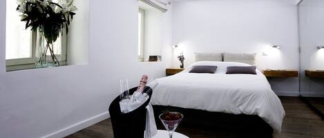 A modern hotel room with a large bed, bedside tables, a small table with a glass of champagne, and a vase of flowers by the window.
