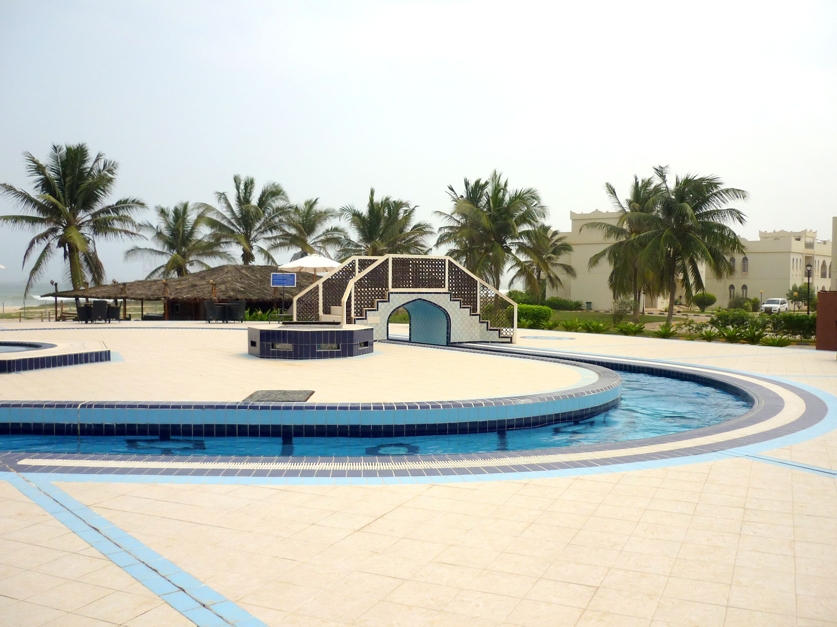 Une piscine dotée d’un îlot central et d’un espace de repos couvert, entourée de palmiers et avec un bâtiment visible au loin.