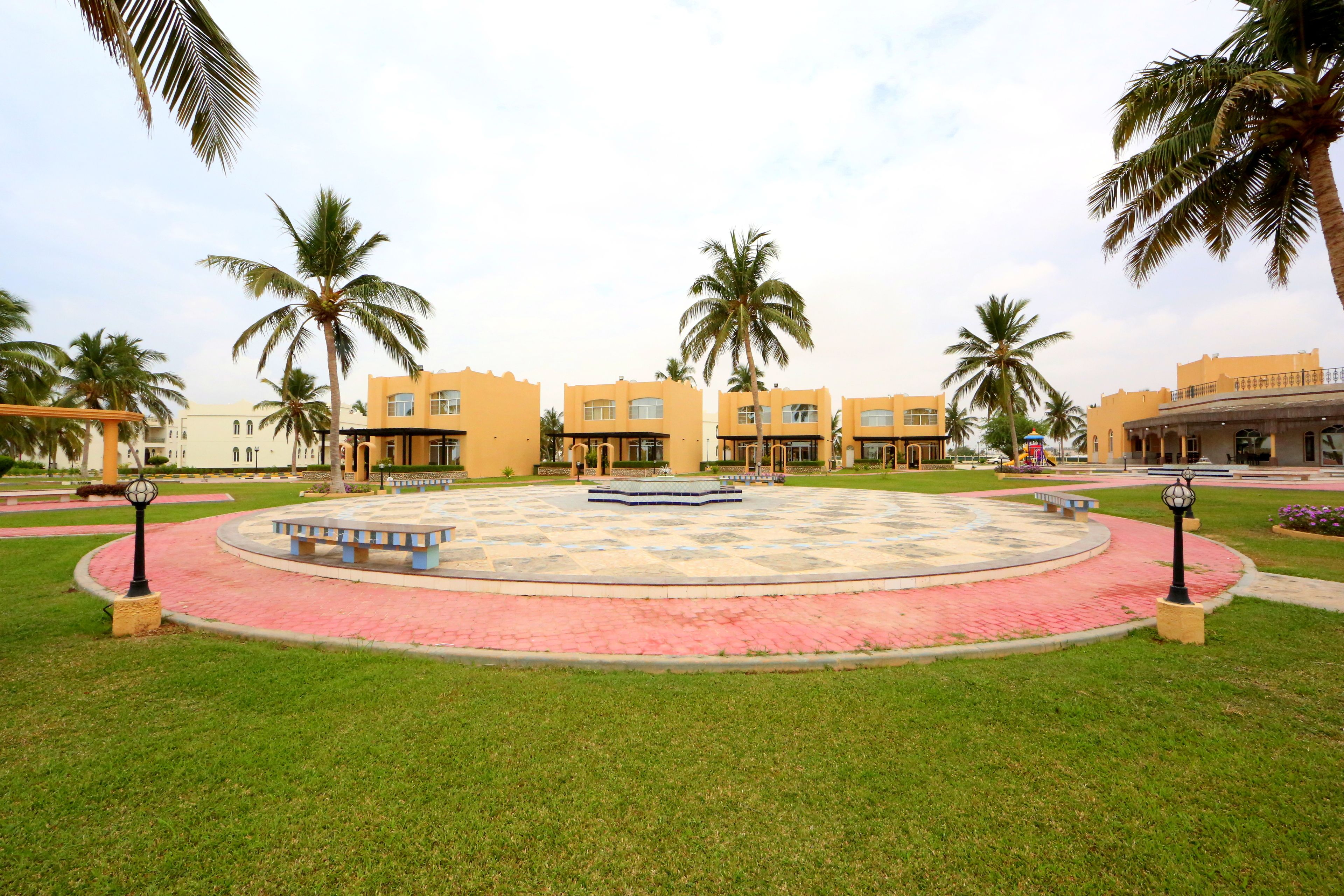 A courtyard with a circular seating area, palm trees, and buildings with yellow facades.