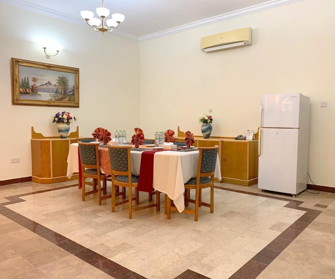 Un coin repas avec une table dressée pour six personnes, entourée de chaises, d’un réfrigérateur et d’un tableau accroché au mur.