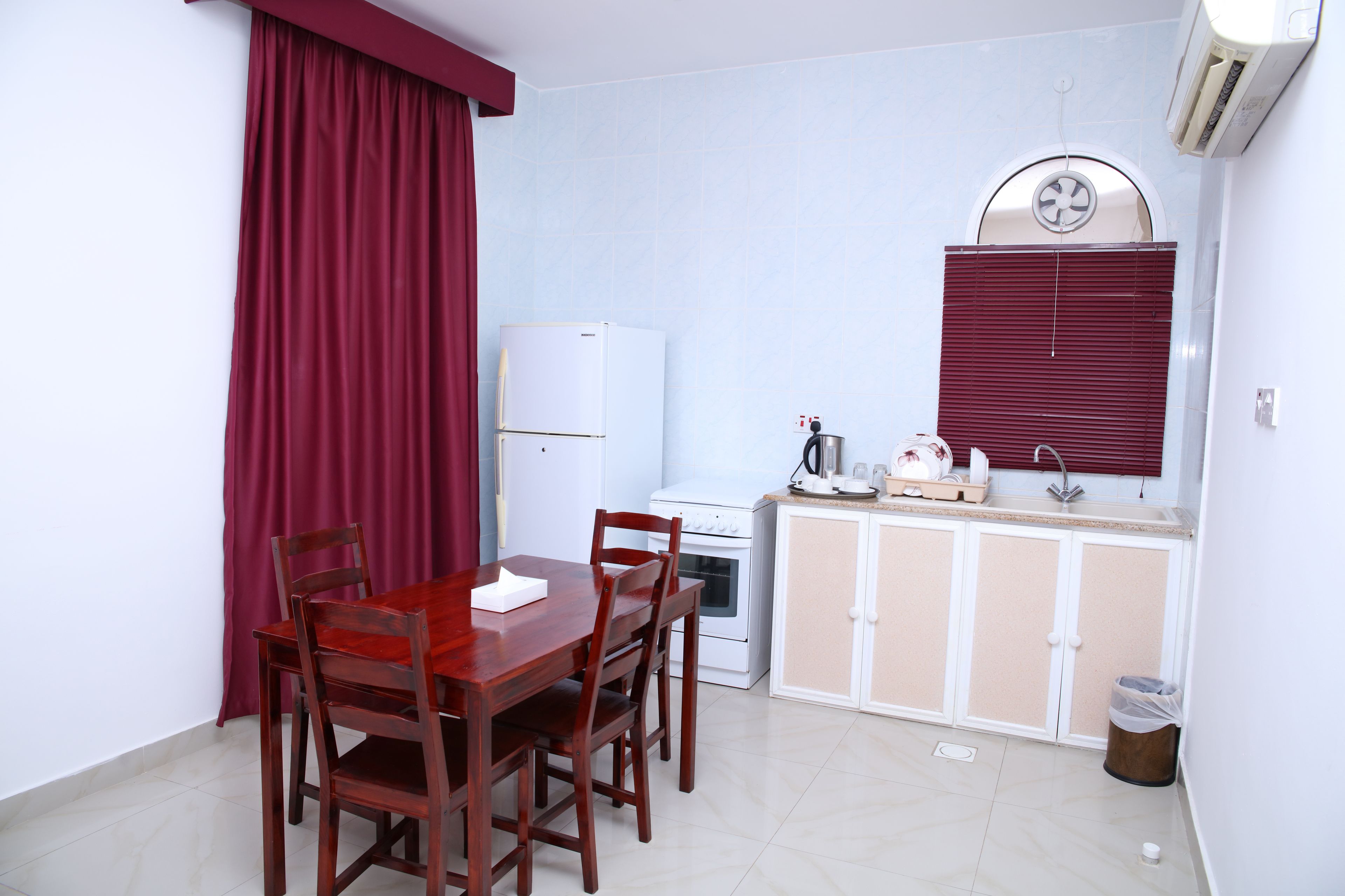 A compact kitchen with a refrigerator, stove, and sink. There is a dining table with chairs and a window with blinds.