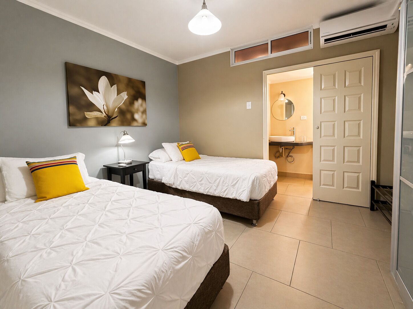 A hotel room with two beds, a nightstand, a lamp, a painting, a bathroom with a sink and mirror, and an air conditioning unit.