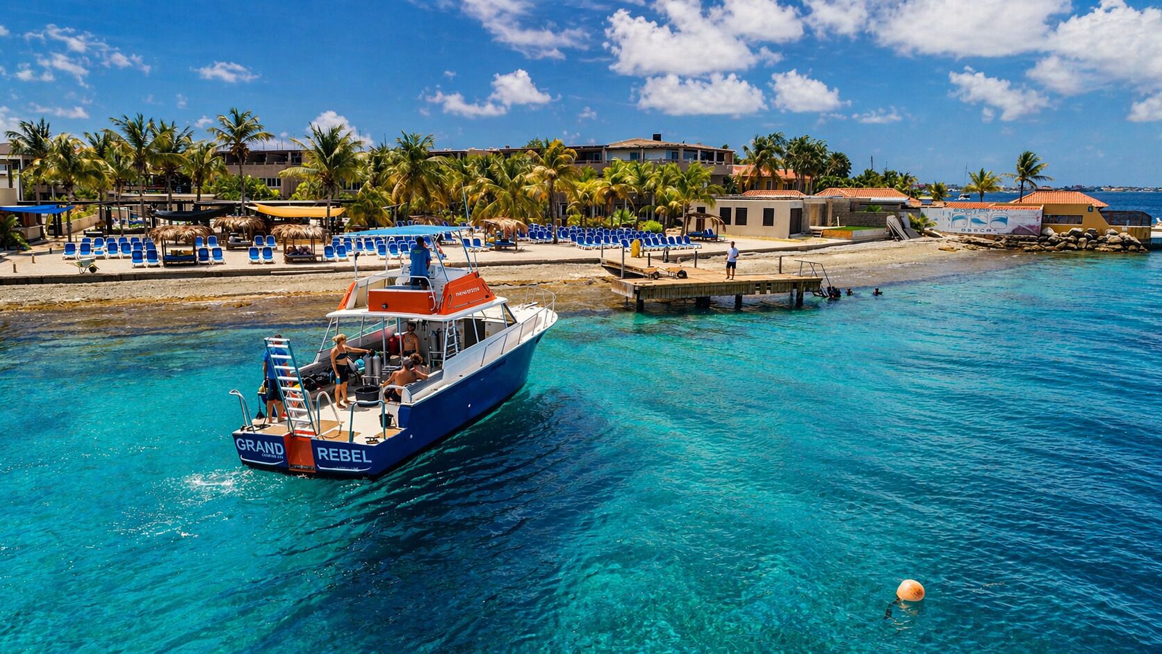 Un barco llamado 'GRAND REBEL' en aguas turquesas cristalinas, una playa con sillas azules y palmeras al fondo.