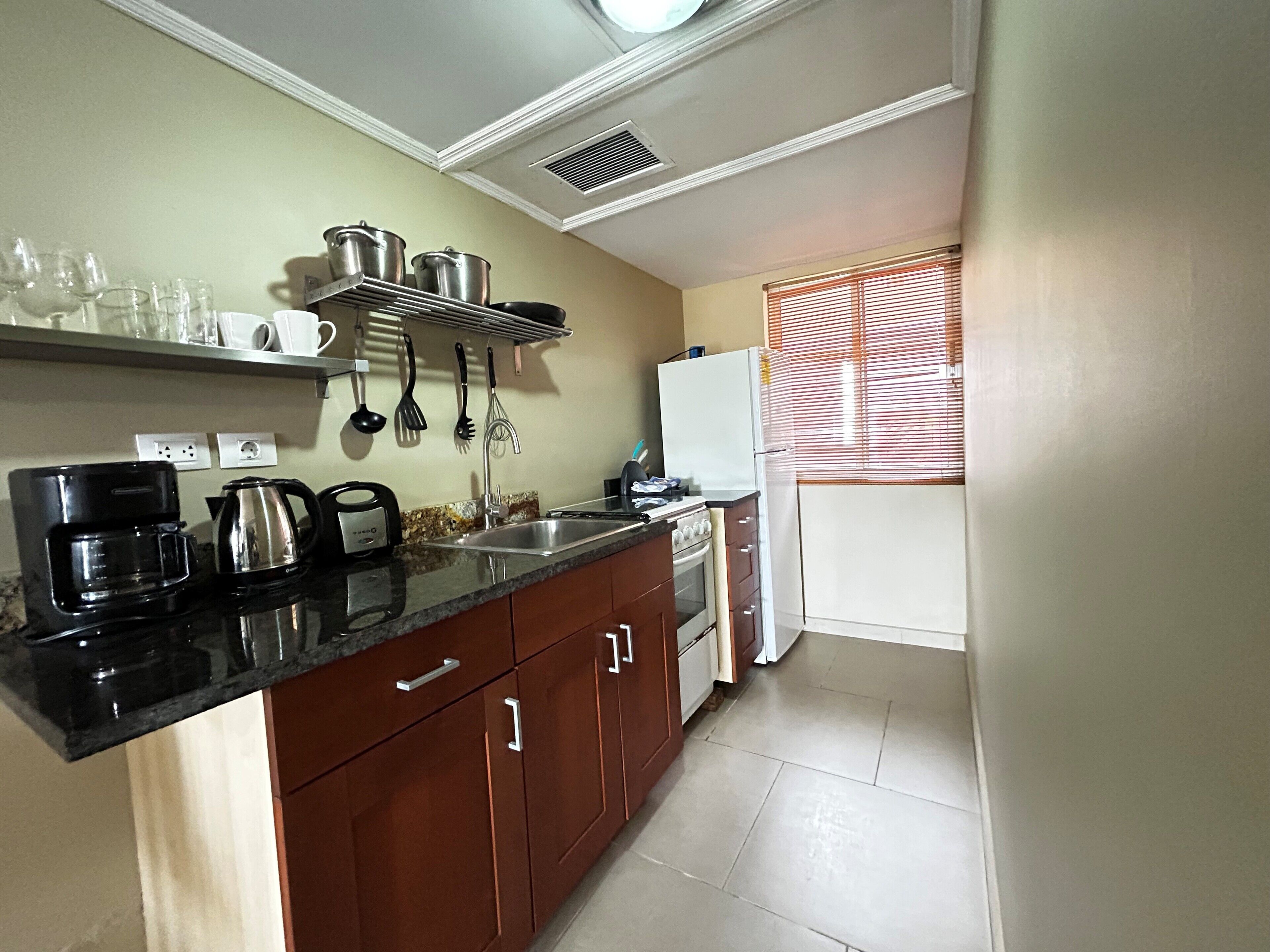 A compact kitchen with a black countertop, stainless steel appliances, and a window with blinds.