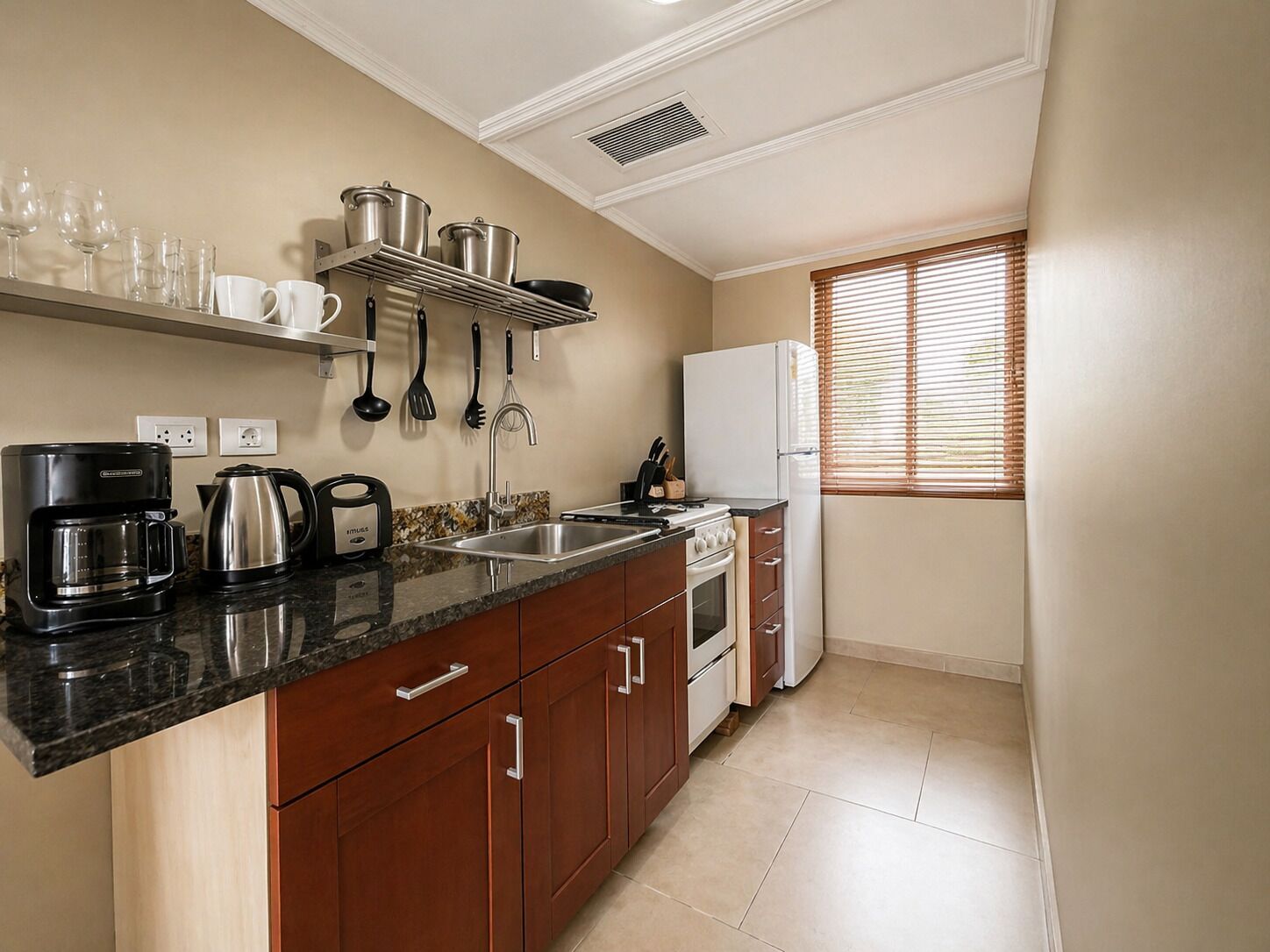 A compact kitchen with a countertop coffee maker, a kettle, a sink, and a stove. There are cabinets with silver handles, a window with blinds, and a wall-mounted shelf with pots and pans.