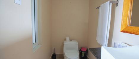A small bathroom with a toilet, a trash bin, a window, and a sink.