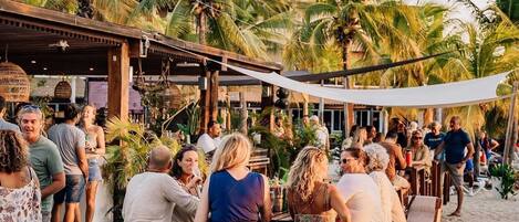 Un espace extérieur aménagé avec des tables et des chaises en bois, situé au bord de la plage, entouré de palmiers et abrité par une structure couverte.