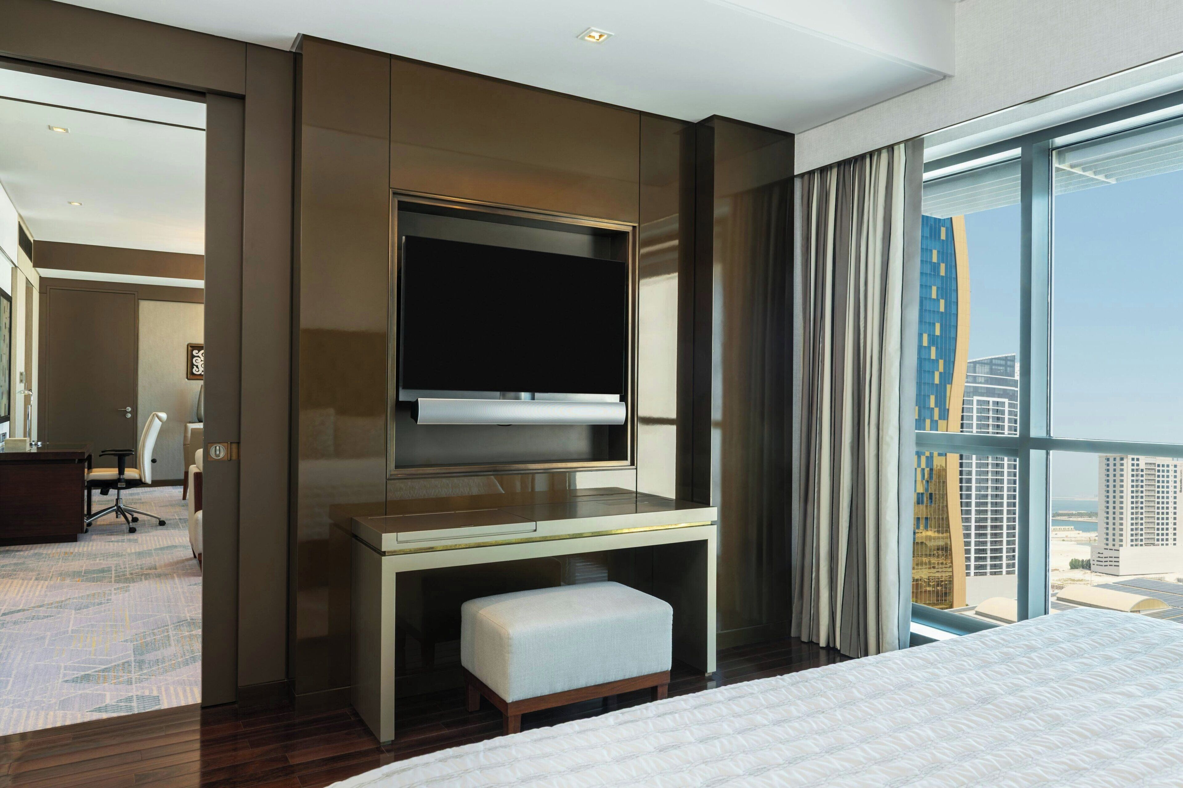Ein modernes Hotelzimmer mit einem großen Bett, einem Schreibtisch mit Fernseher, einer kleinen Sitzecke und einem großen Fenster mit Blick auf die Stadt.