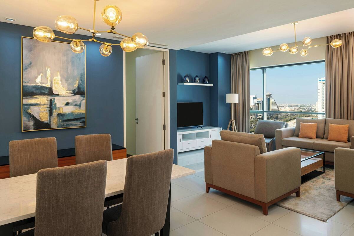 A modern living room with a dining area, a large painting, a flat-screen TV, and a city view through large windows.