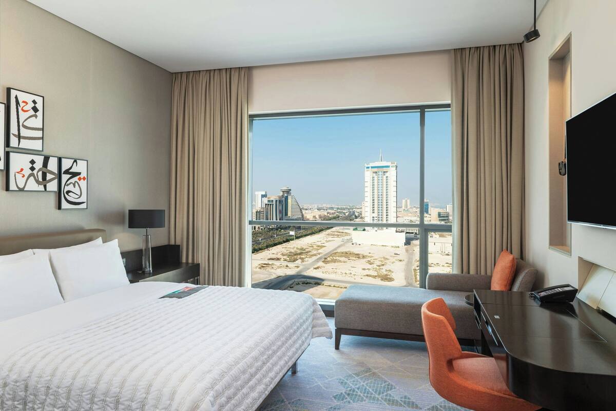 A hotel room with a large bed, a desk, a chair, and a view of the cityscape through a large window.