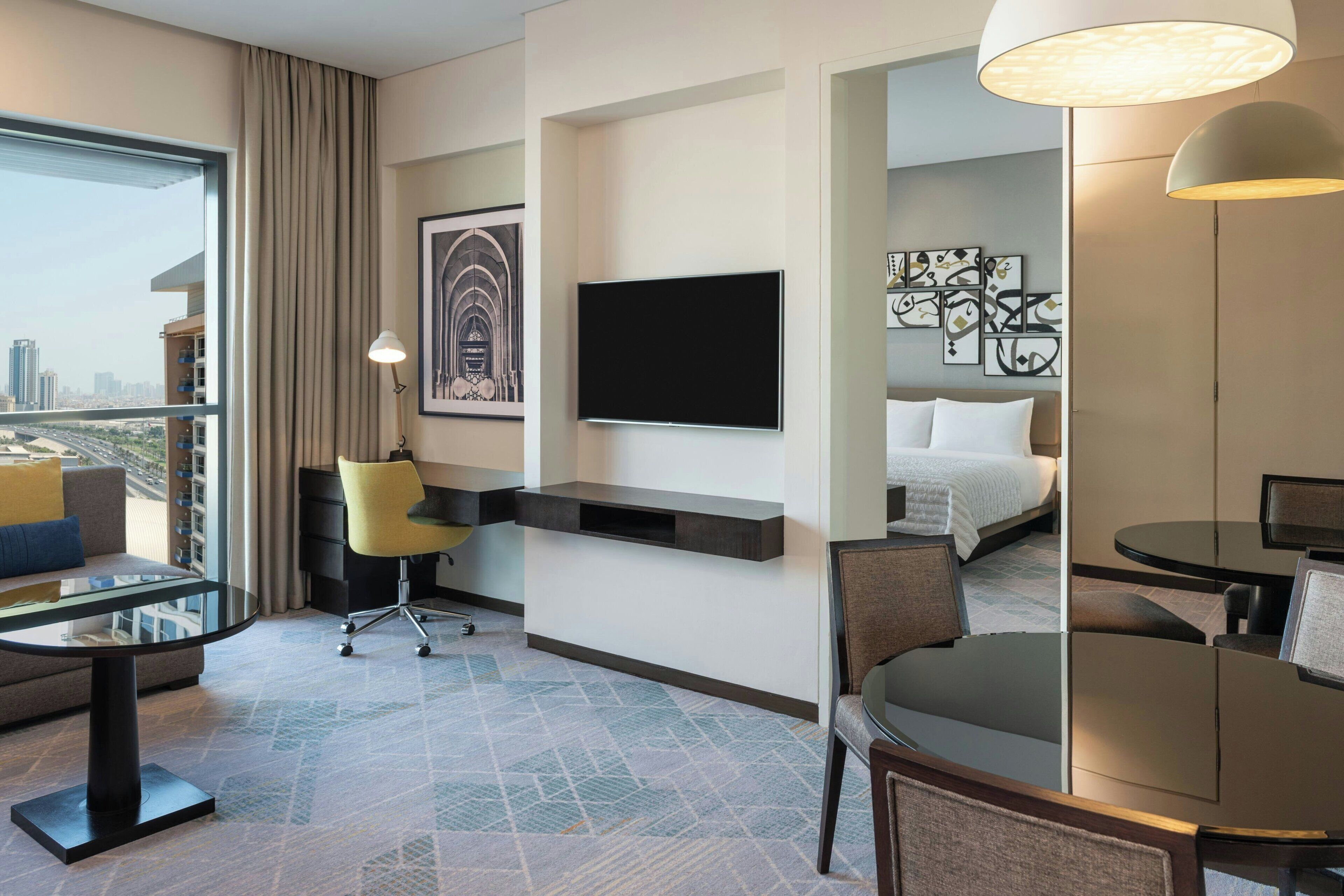 Ein Hotelzimmer mit großem Fenster, einem Flachbildfernseher, einem Schreibtisch mit Stuhl, einem Bett und einer Essecke.