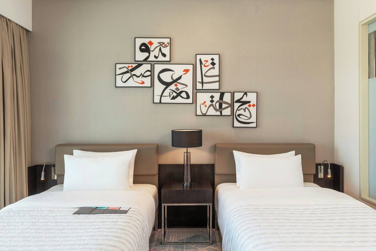 A hotel room with two beds, a nightstand with a lamp, and artwork on the wall.