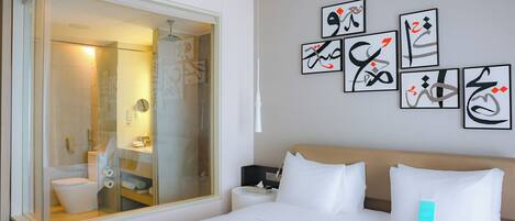 Habitación de hotel con una cama grande, una mesita de noche y un baño con cabina de ducha de vidrio, además de una obra de arte en la pared.