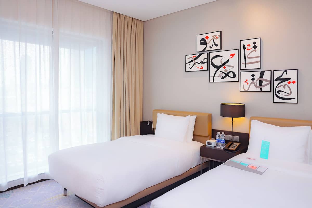 A hotel room with two beds, a nightstand with a lamp, a phone, and artwork on the wall.