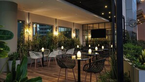 Un área de comedor moderna al aire libre con mesas y sillas redondas, rodeada de plantas en macetas y un piso de madera.