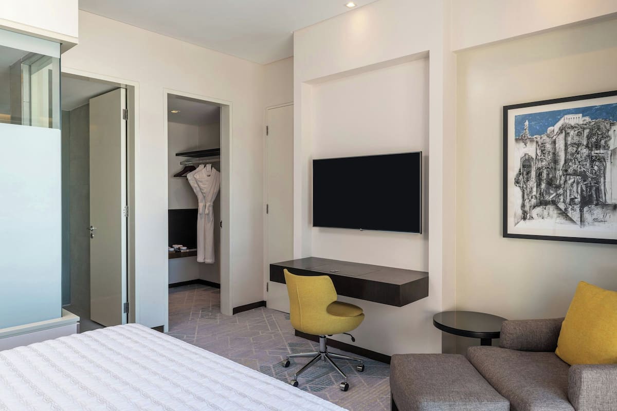 A modern hotel room with a bed, a desk with a chair, a flat-screen TV, and a painting on the wall.