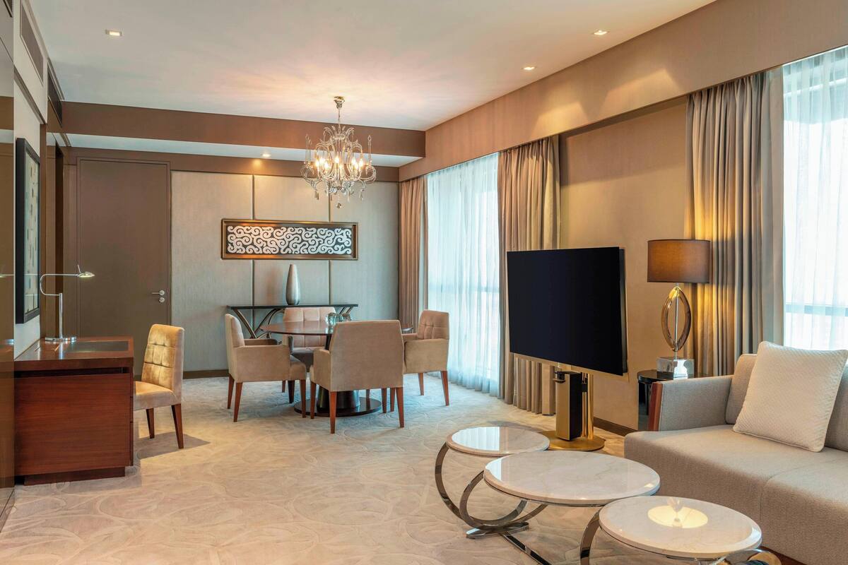 A modern hotel room with a dining area, a sofa, a TV, and a chandelier.