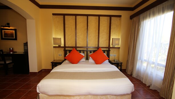 A large bed with two orange pillows, a nightstand with a lamp, and a desk with a chair in a hotel room. - Tamra Beach Resort (Sharm El Sheikh)