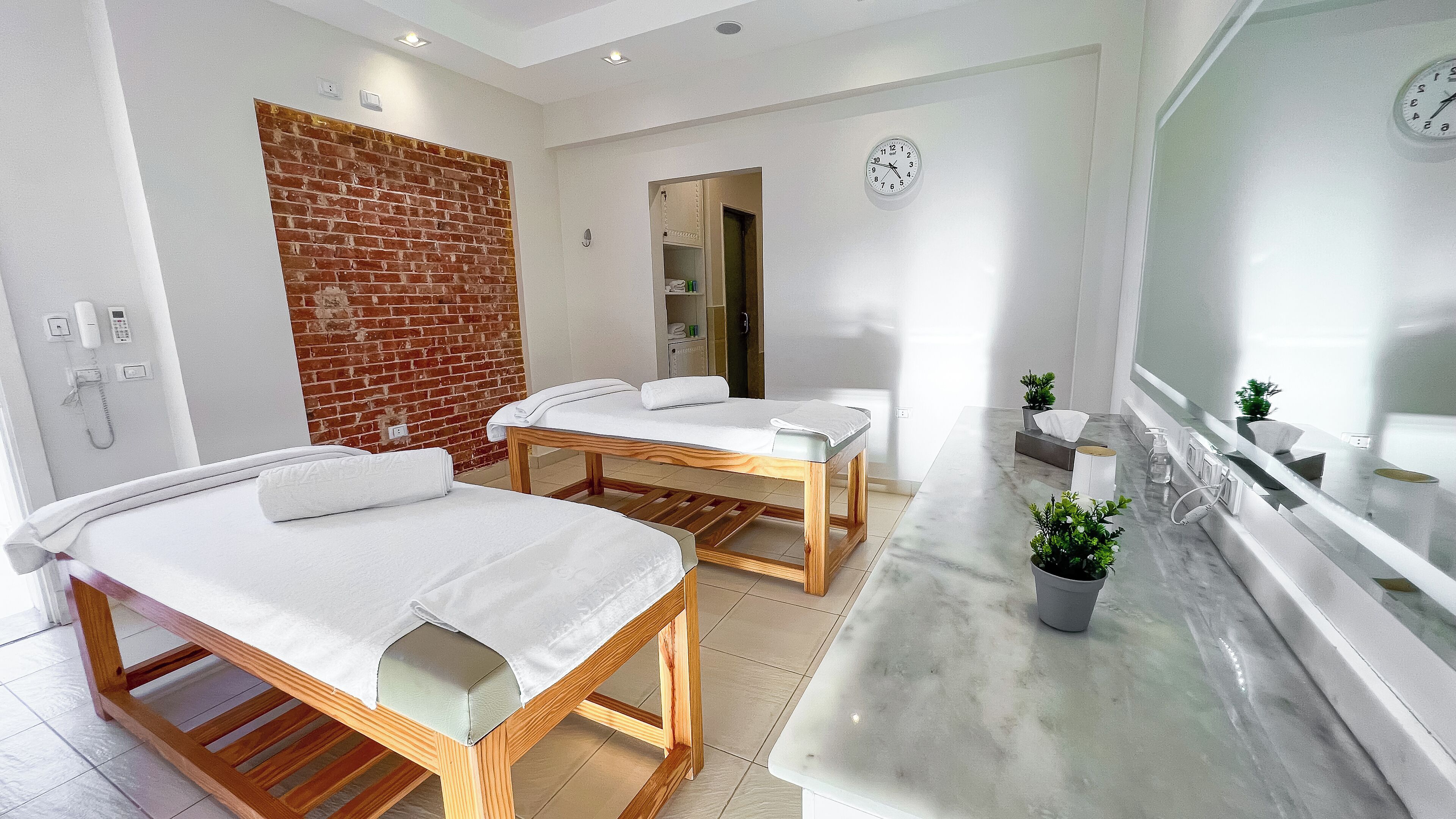 Un cuarto de spa con dos camillas de madera, un mostrador de mármol, una pared de ladrillo y un reloj en la pared.