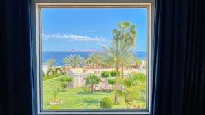 Une vue depuis une fenêtre sur un jardin bien entretenu, avec des palmiers, une pelouse et, au loin, la mer et les montagnes.