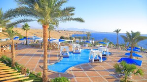 Un espace piscine offrant une vue dégagée sur la mer, entouré de palmiers et agrémenté de parasols bleus.