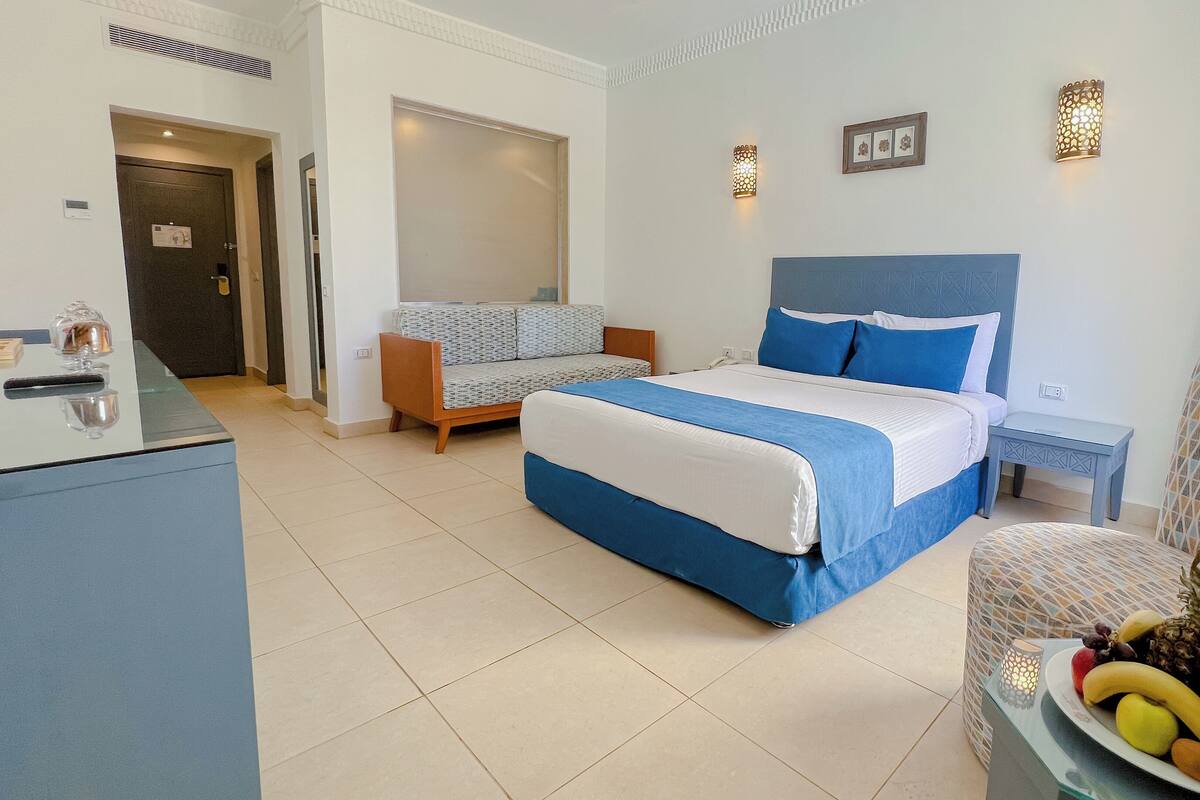 A hotel room with a bed, a sofa, a chair, a small table with fruit, and a desk with a lamp.