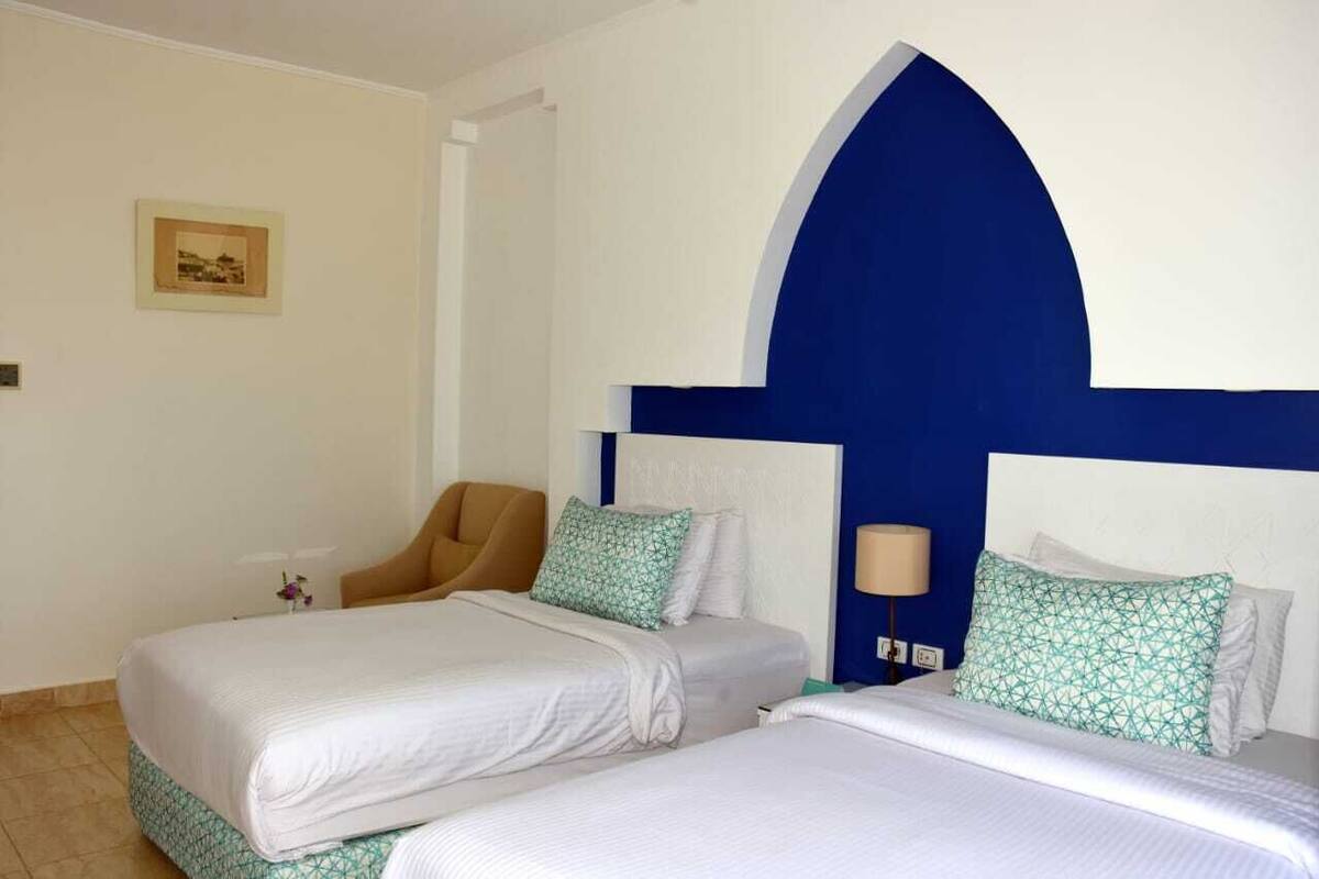 A hotel room with two beds, a chair, and a wall-mounted lamp.