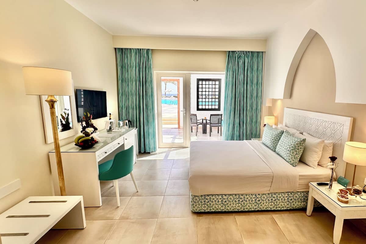 A hotel room with a bed, bedside table, desk, and a view of the pool area.