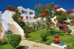 Ein weißes Gebäude mit einer Treppe, die zu einer Tür führt, umgeben von einem gepflegten Garten mit verschiedenen Pflanzen und einem großen, roten Blumentopf.