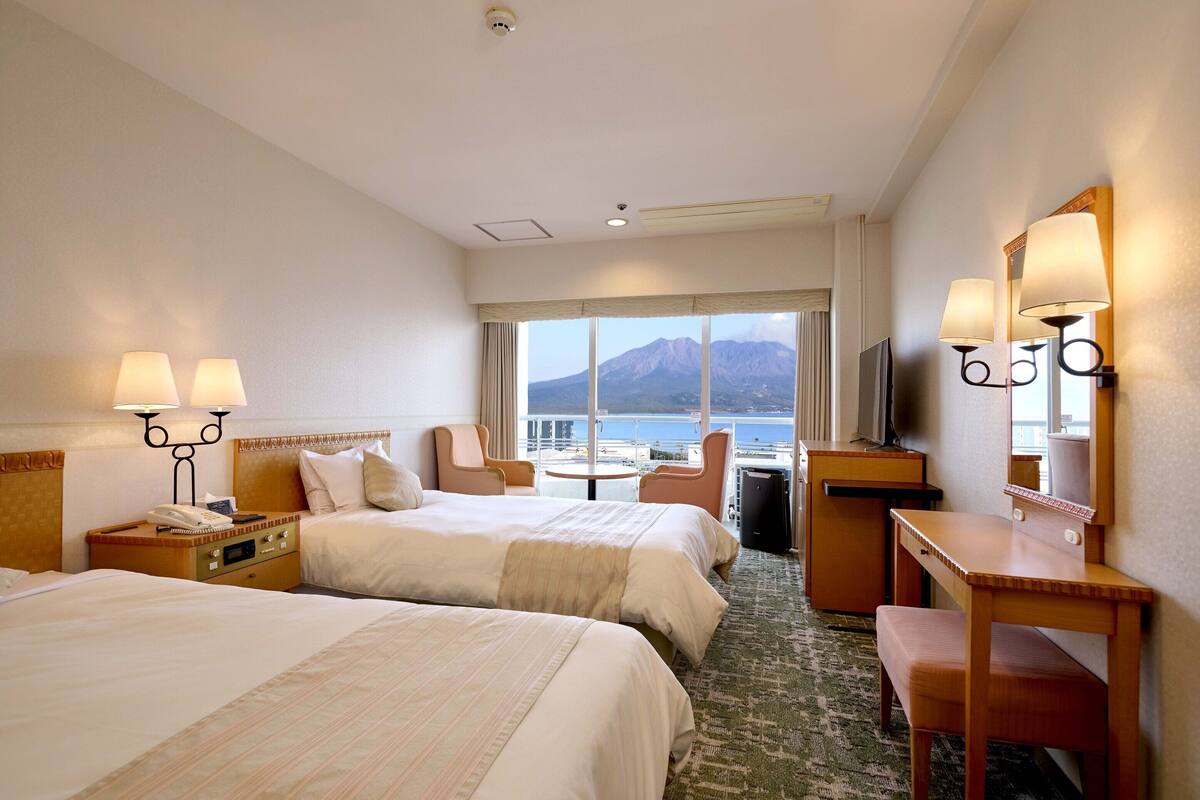 Habitación de hotel con dos camas, un escritorio, una silla, un televisor y vistas al mar y a las montañas.