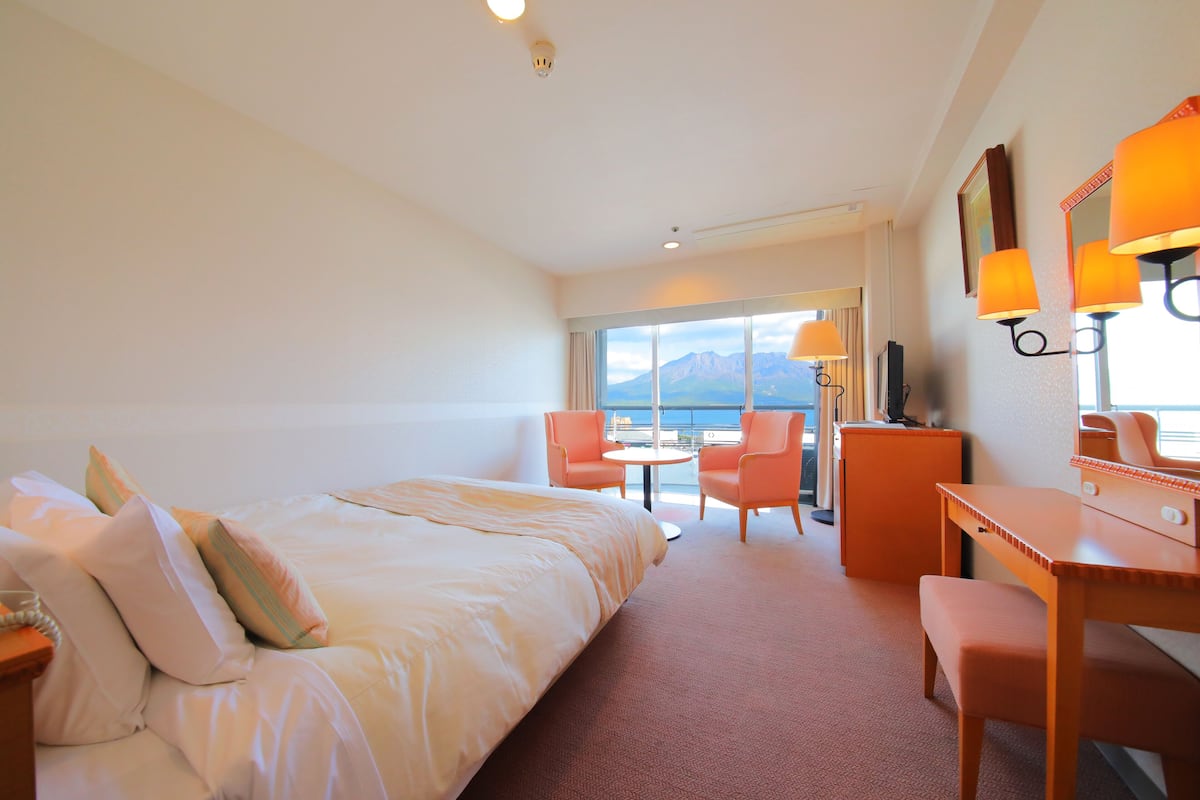 Habitación de hotel con cama, escritorio, dos sillas, una mesita y un ventanal grande con vistas a montañas.