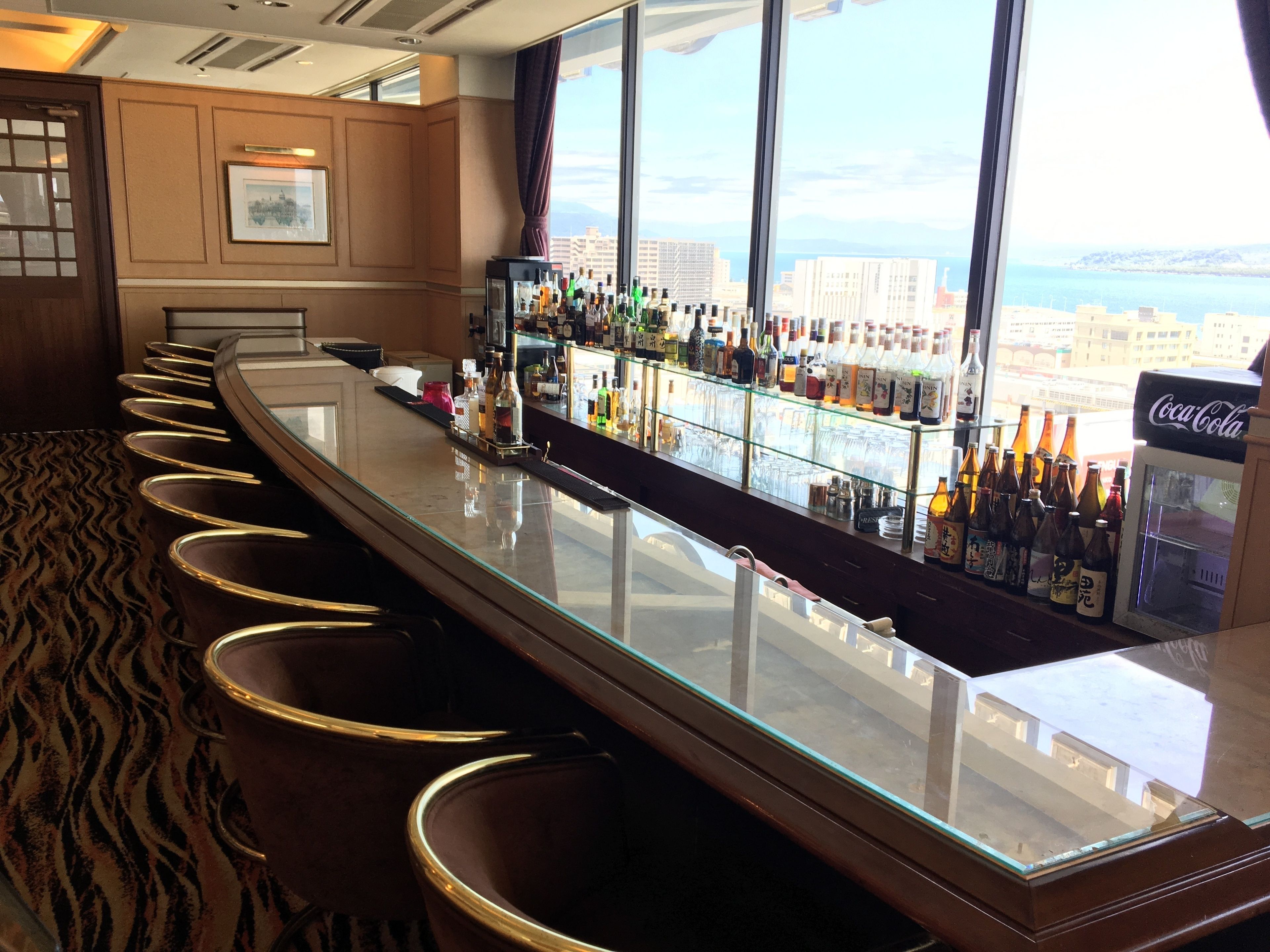 A bar with a long counter and multiple stools, a glass display case with bottles, and a large window offering a view of buildings and a body of water.