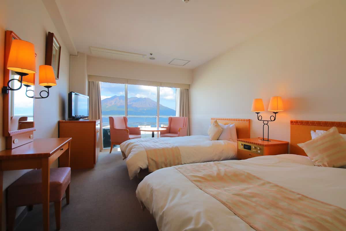 Habitación de hotel con dos camas, un escritorio, una silla y un televisor. Se ve una ventana con vista a las montañas.