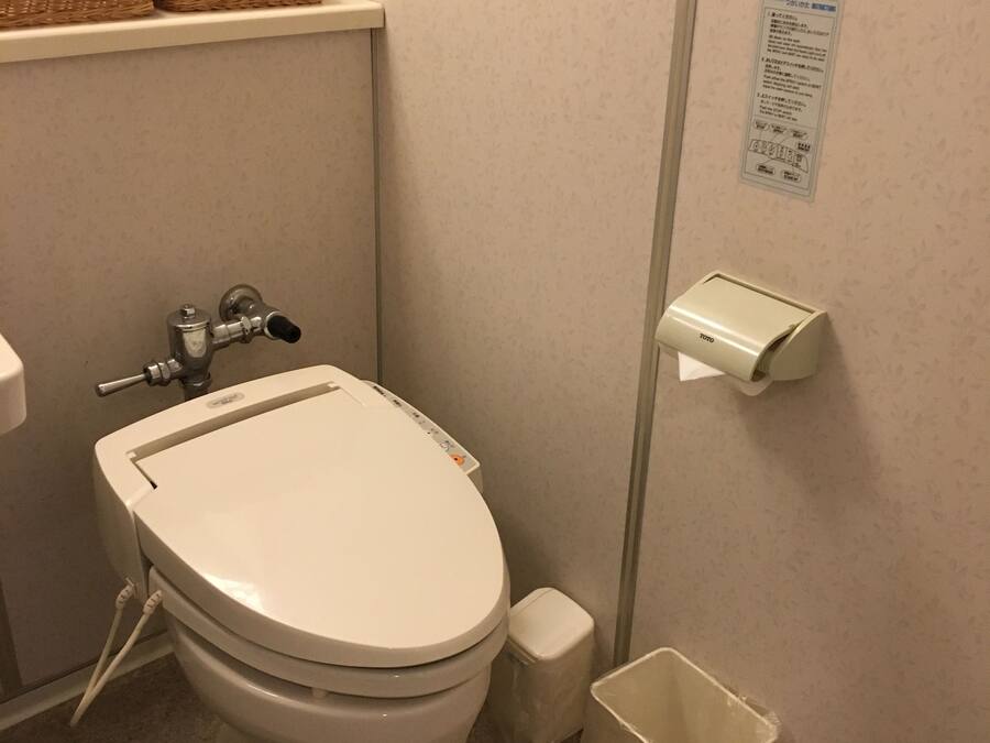 Un baño con inodoro, un cubo de basura, dispensador de toallas de papel y un estante con cestas.