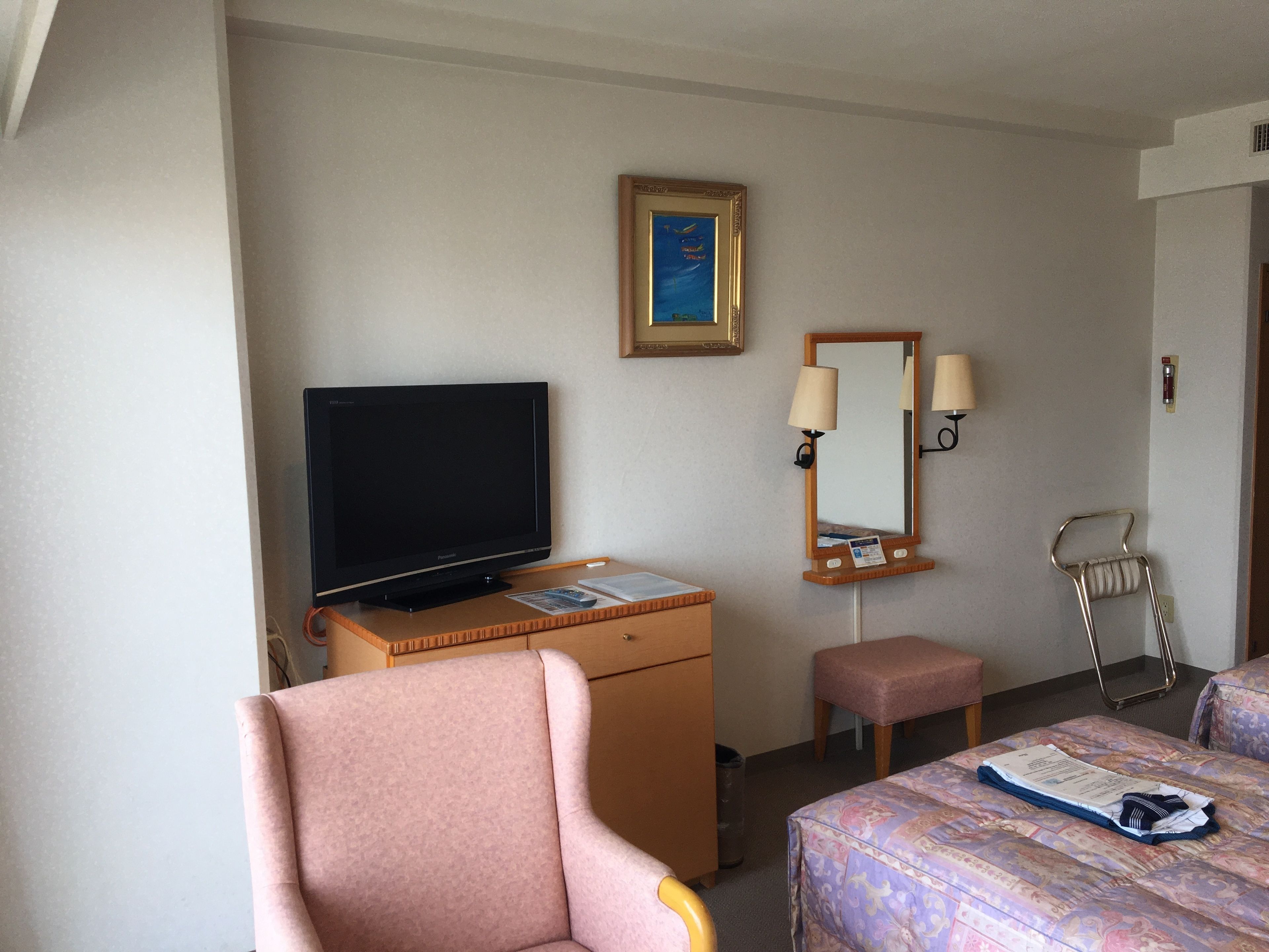 A hotel room with a television on a wooden dresser, a mirror, a lamp, a chair, and a bed with a floral bedspread.