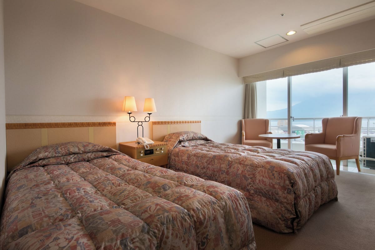 Habitación de hotel con dos camas, una mesita de noche con lámpara, una silla y un balcón con vistas.