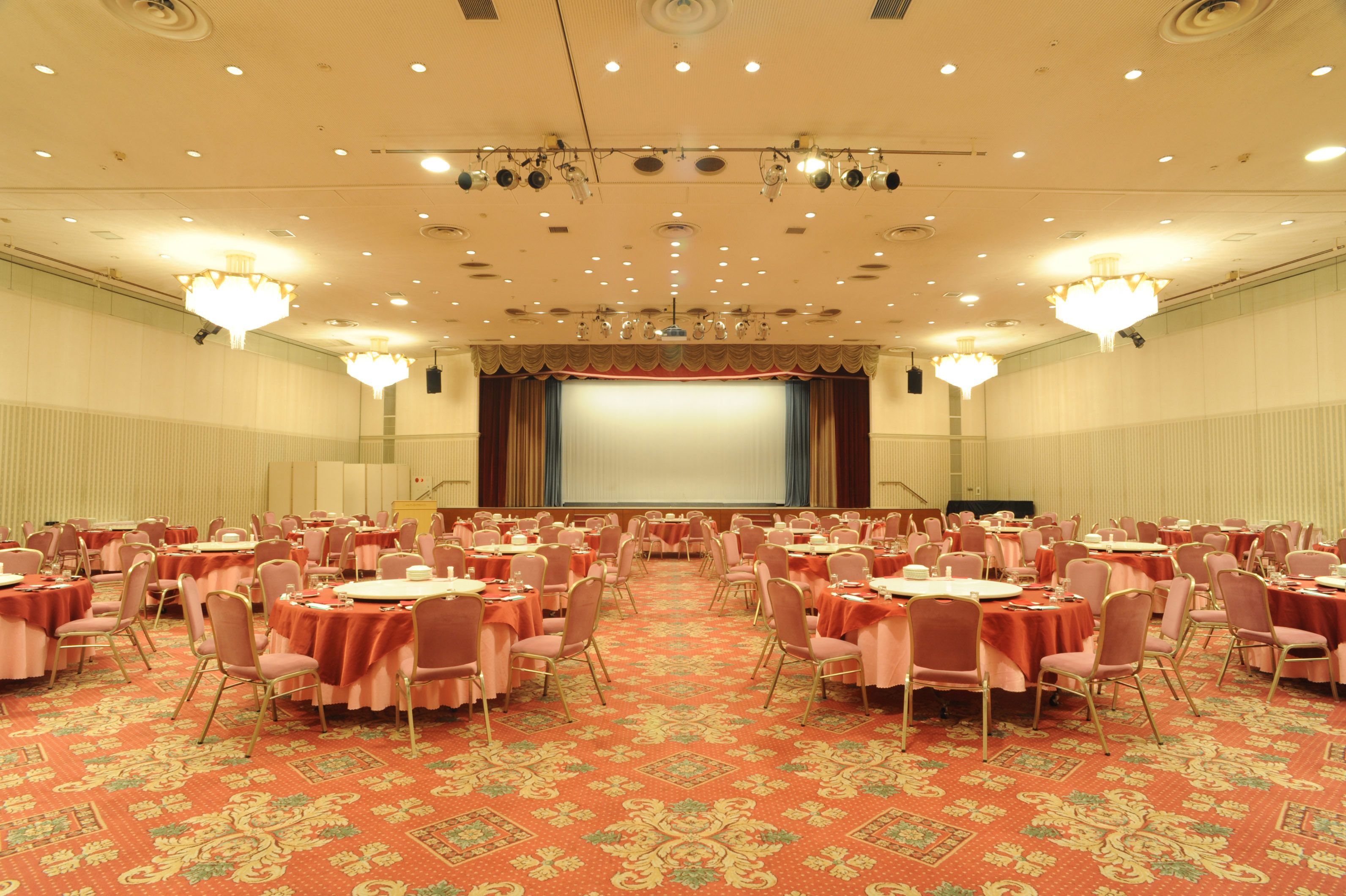 A large conference room with round tables and chairs, a stage with a screen, and chandeliers.