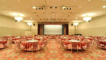 A large conference room with round tables and chairs, a stage with a screen, and chandeliers.