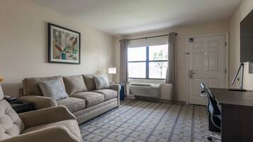 A living room with a beige sofa, a small table, a radiator, a window with curtains, and a door.