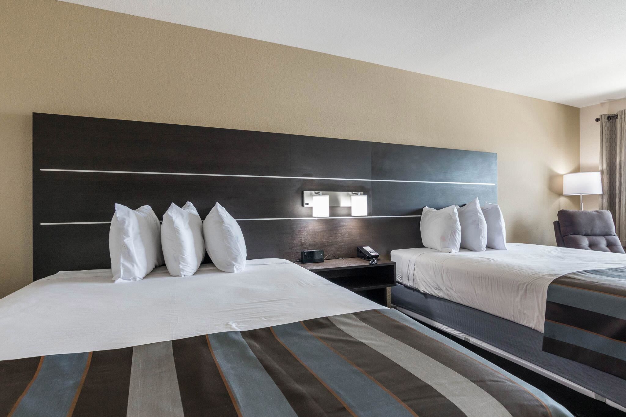 A hotel room with two beds, a large dark headboard, bedside tables, a lamp, and a view of the room.