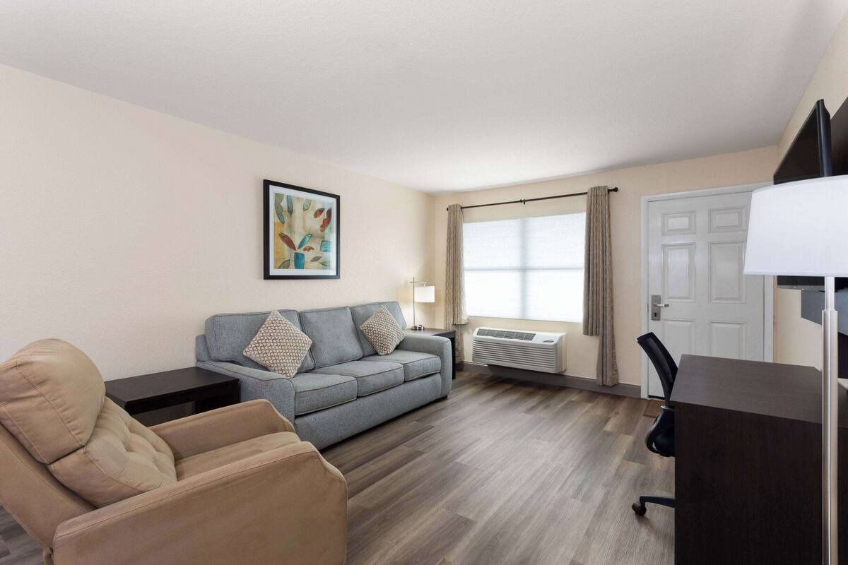 Une chambre d’hôtel moderne avec un canapé gris, un fauteuil beige, un bureau avec un ordinateur et une grande fenêtre avec des rideaux.