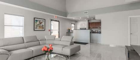 A spacious living area with a grey sectional sofa, a glass coffee table with flowers, and a kitchen in the background.