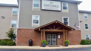 A Stay Lodge Extended Stay building with a brick facade, a wooden entrance canopy, and two potted plants at the entrance. - Stay Plus Extended Stay Suites (Auburn)